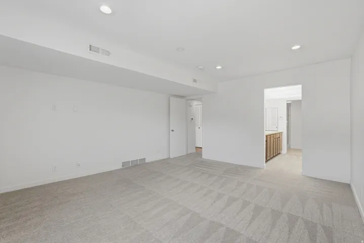 Recessed ceiling lighting illuminates a spacious room featuring light-colored patterned carpeting and crisp white walls