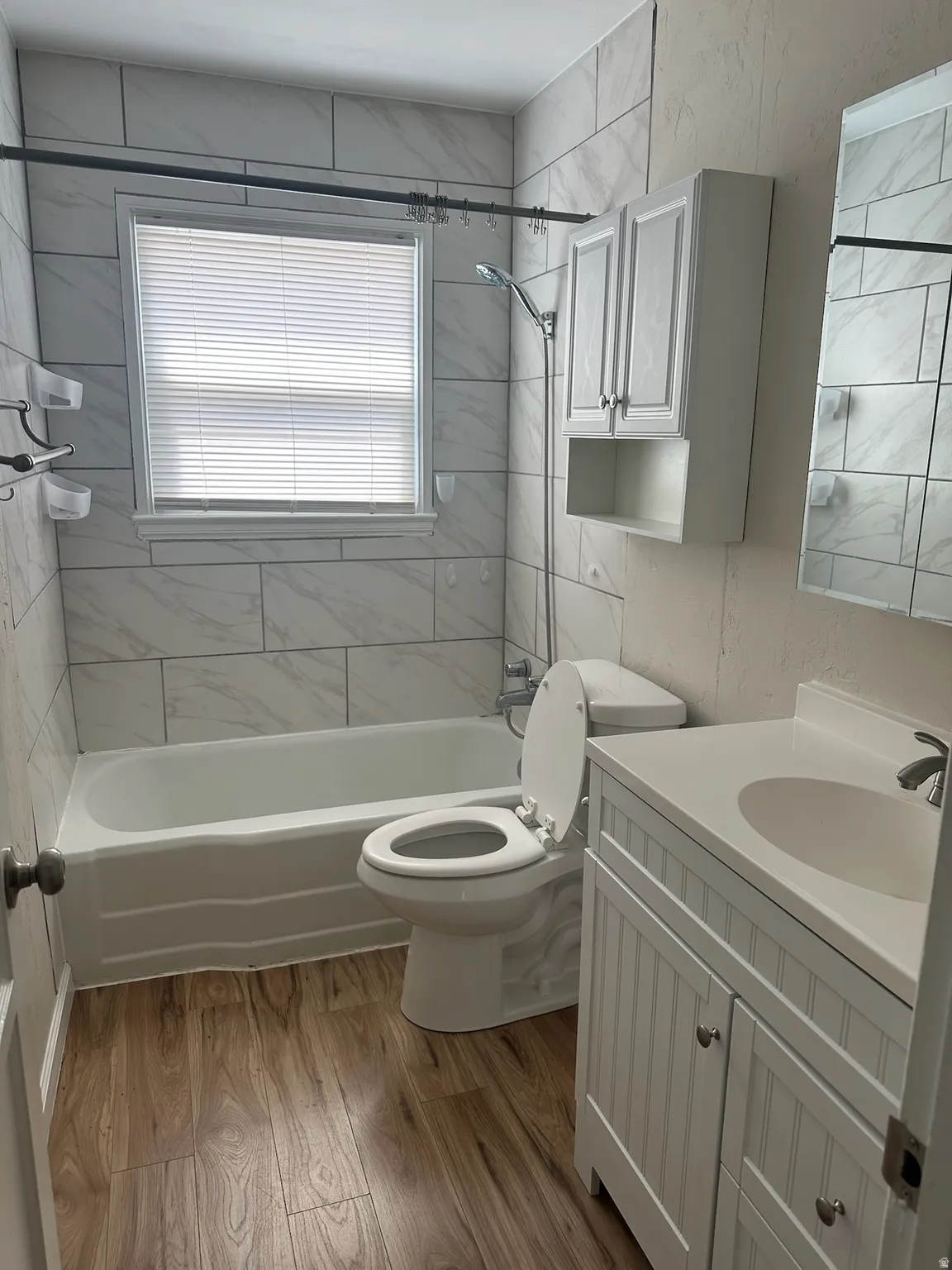 Bathroom featuring wood-finish flooring, white tiled shower surround, bathtub, white vanity with integrated sink, and white wall-mounted cabinet
