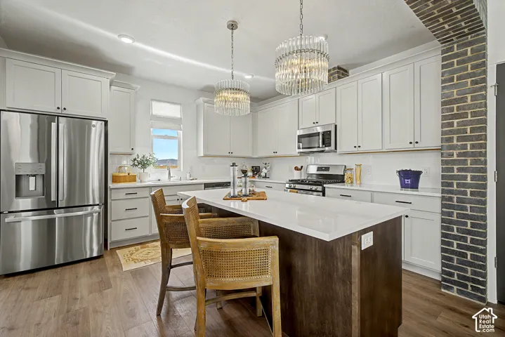 White shaker cabinetry, stainless steel appliances, central island with seating, wood-finish flooring, and brick architectural detail