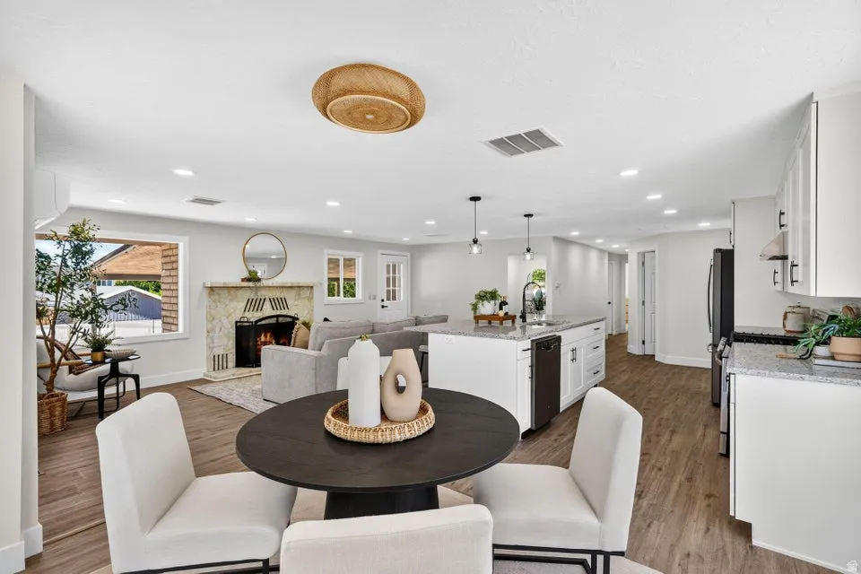 Open-concept living area featuring wood-finish flooring, a stone-faced fireplace, a kitchen island with a built-in sink, white cabinetry, and recessed lighting