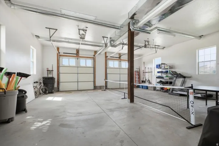 Spacious garage featuring two overhead doors with window accents, exposed wood beam, multiple windows for natural light, and exposed overhead ductwork