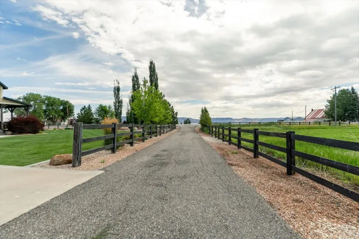Paved driveway with a split-rail fence, manicured lawn, and mature trees