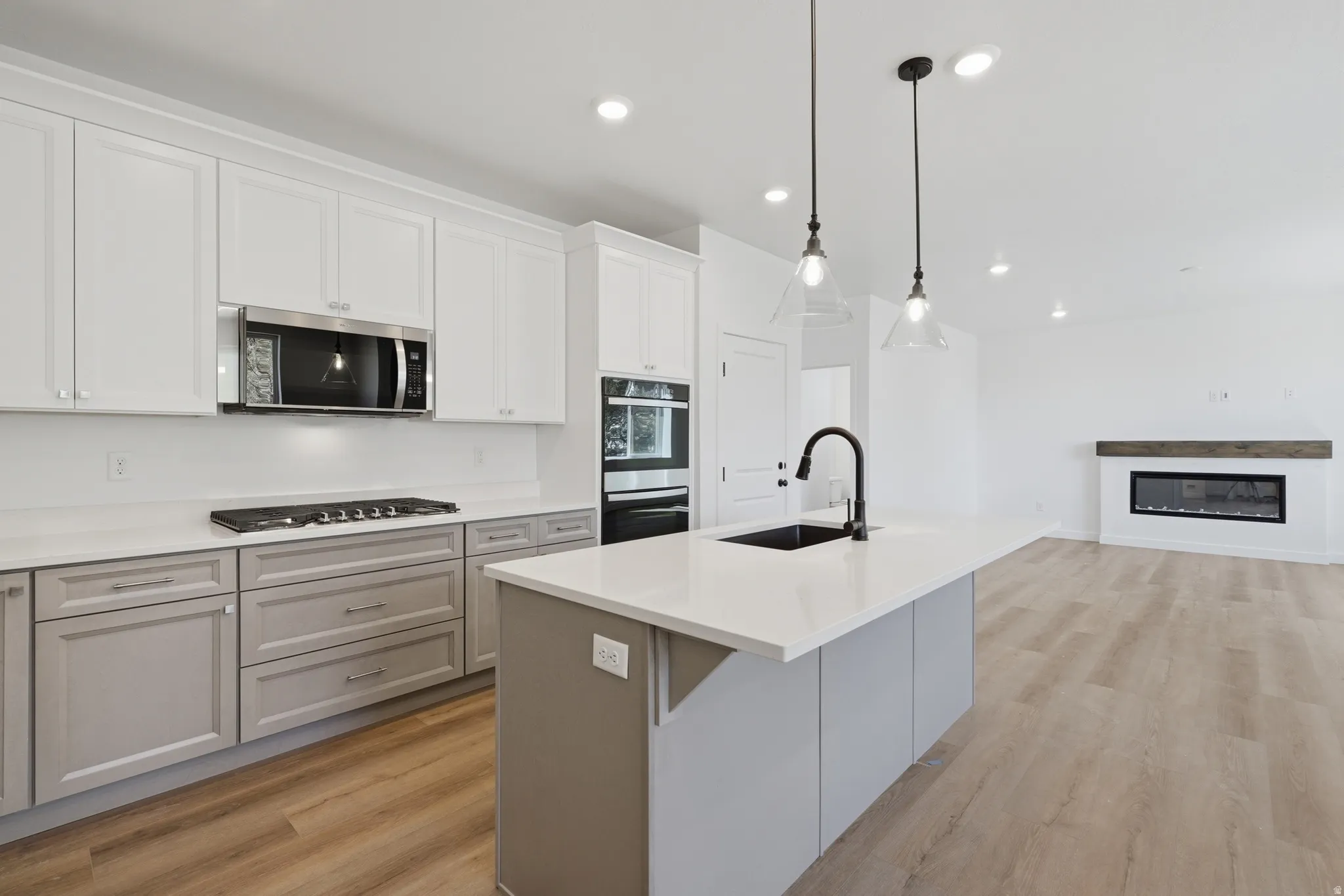 Kitchen with two-tone cabinetry, stainless steel appliances, a large island with a sink, pendant lighting, and wood-finish flooring extending into the living area with a fireplace