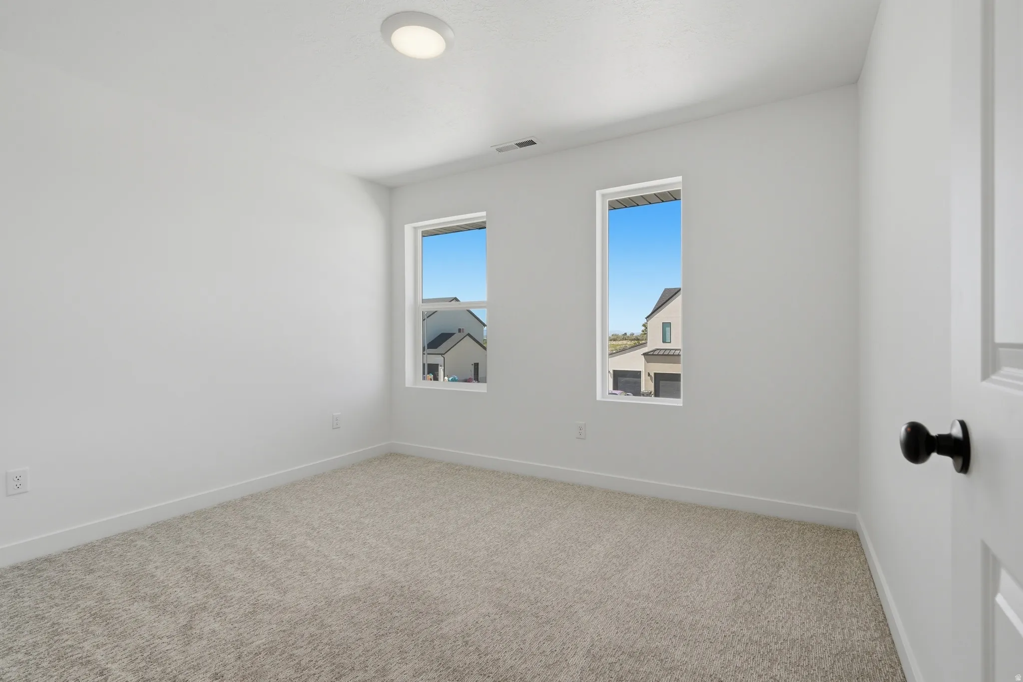Recessed ceiling light fixture, two vertical windows, light neutral carpeting, white interior walls, and a white door with a black doorknob