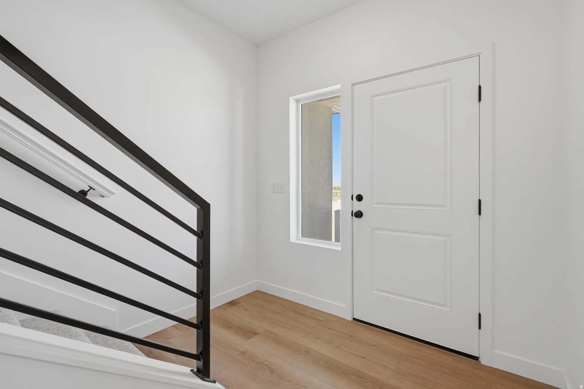 Wood-finish flooring, white paneled door, black hardware, black railing, and a window providing natural illumination