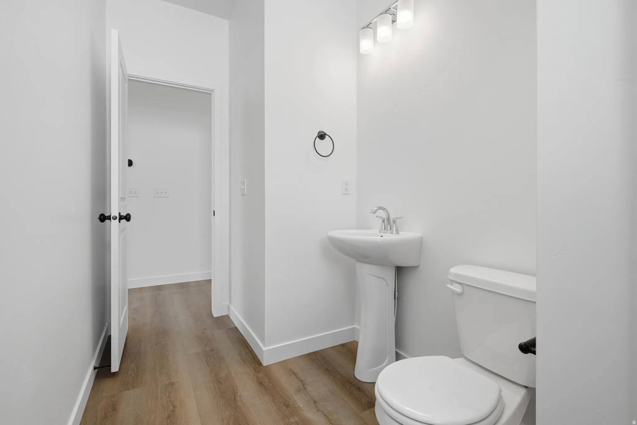Pedestal sink, wall-mounted light fixture, wood-finish flooring, white walls, and a white toilet