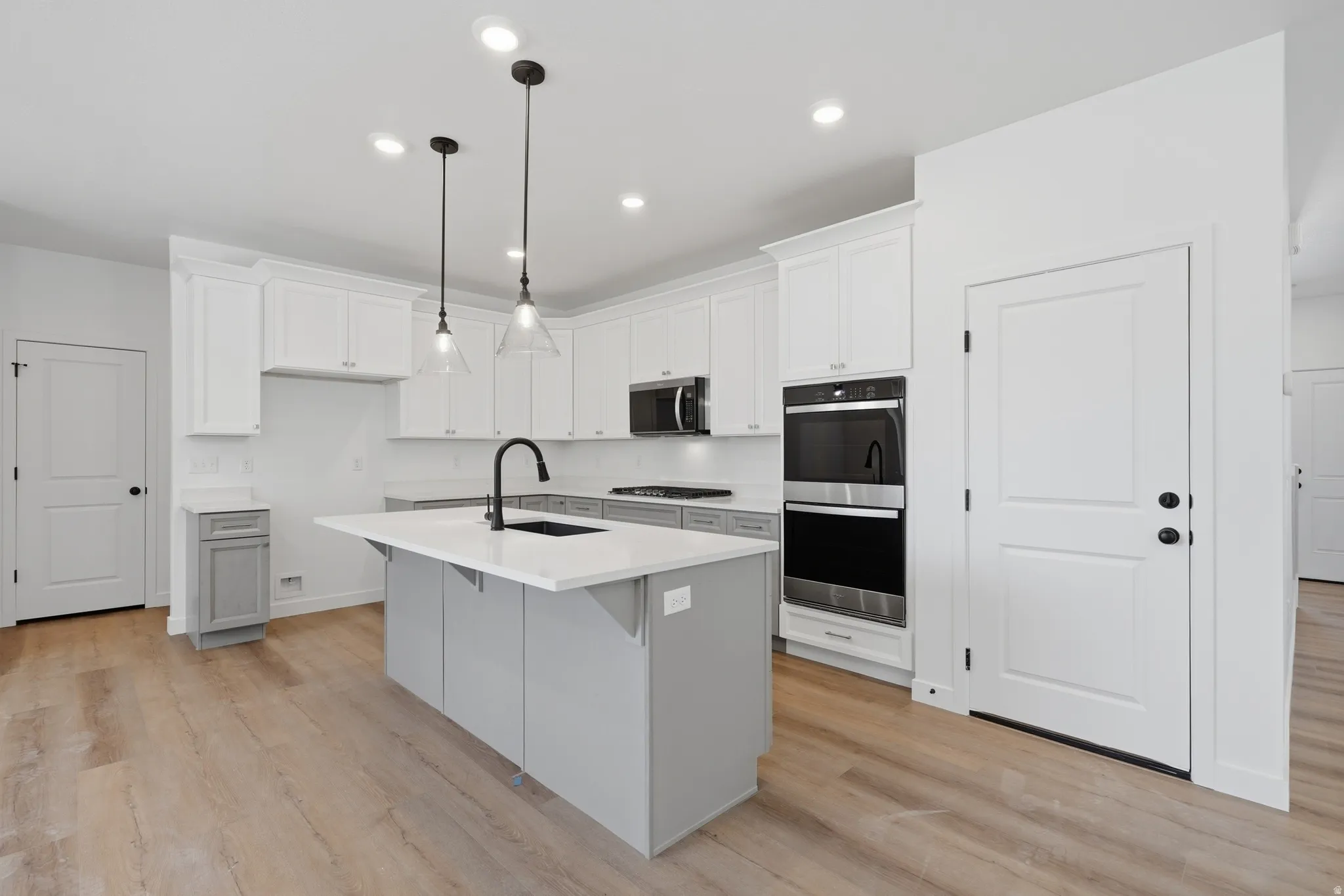 Kitchen featuring light wood-finish flooring, a central island with a white countertop and dark sink, pendant lighting, white upper cabinetry, and integrated dark appliances