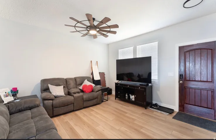 Light-toned wood-finish flooring, two large windows with blinds, a dark wood entry door, and a multi-blade ceiling fan