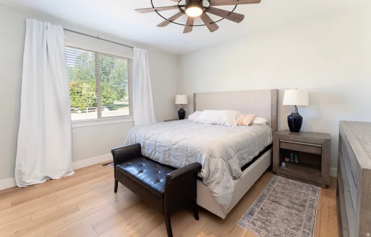 Light wood-finish flooring, a ceiling fan with integrated lighting, and a window with blinds and white curtains