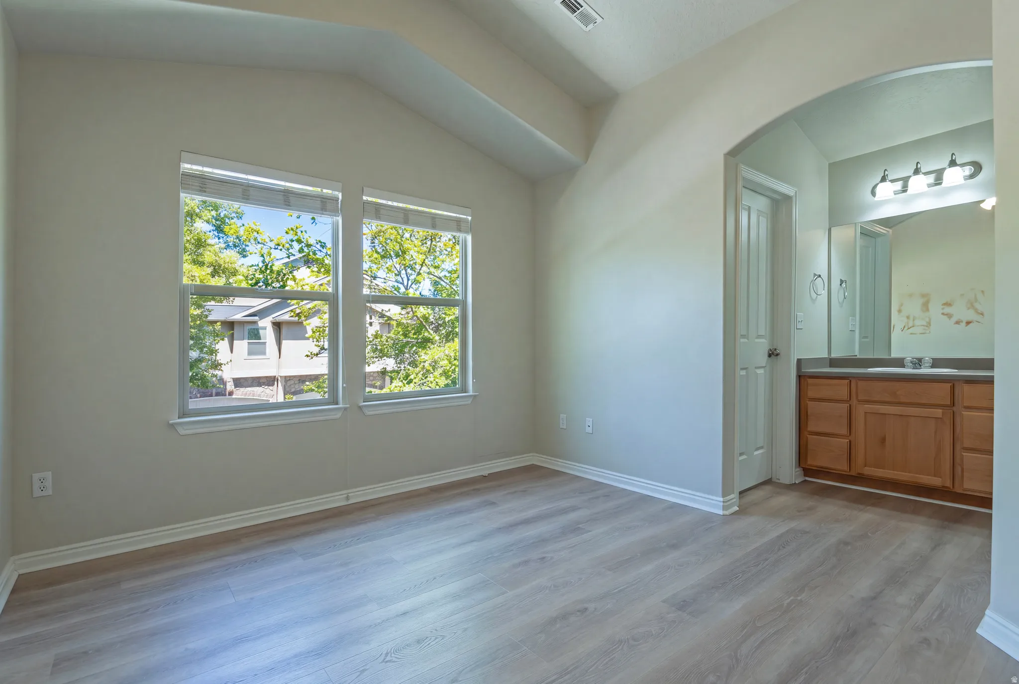 Vaulted ceiling, dual windows, wood-finish flooring, arched entryway, and a dual-sink vanity with wood cabinetry