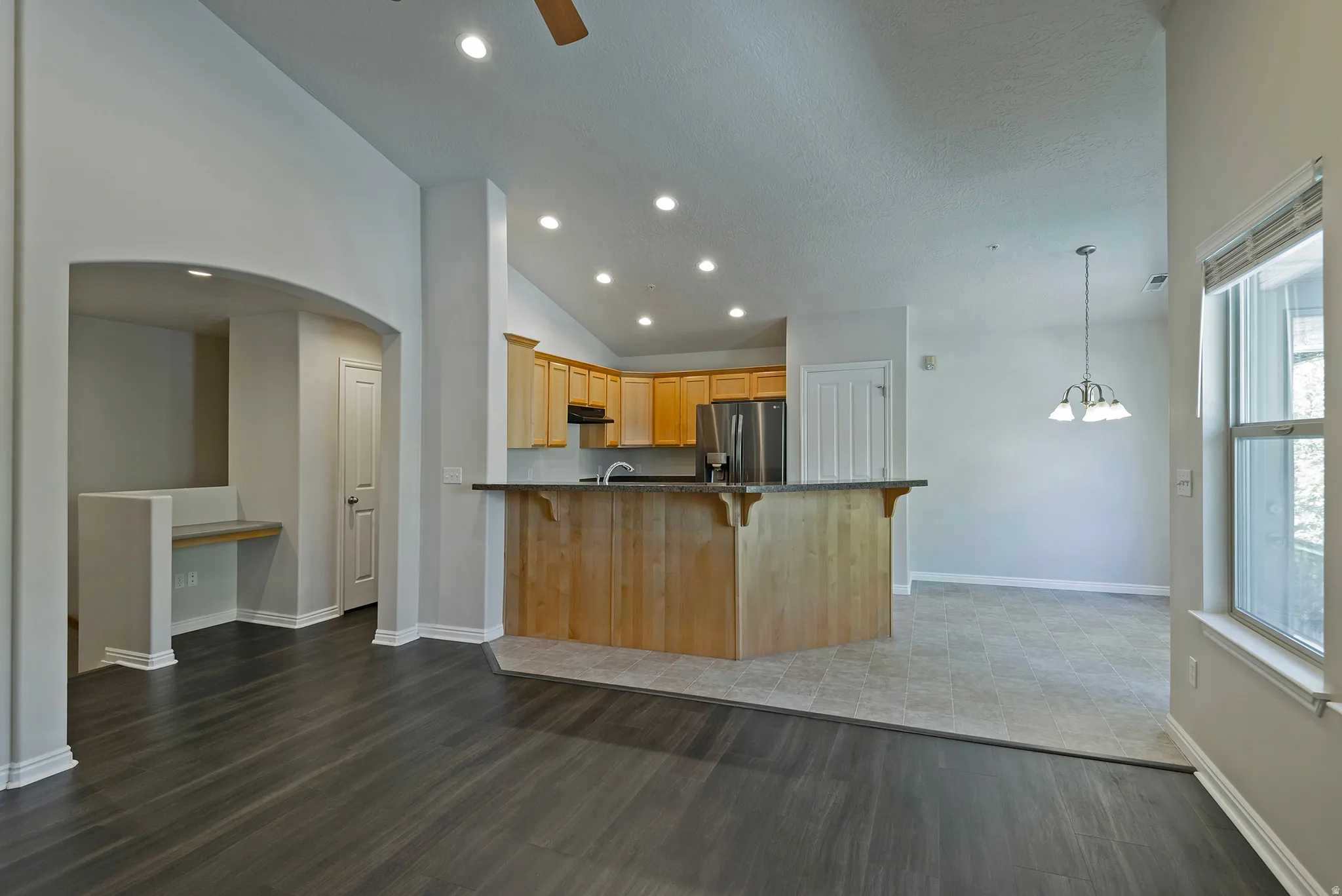 Open-concept floor plan featuring wood-finish flooring, vaulted ceilings with recessed lighting, a kitchen with wood cabinetry and stainless steel appliances, and a built-in desk area