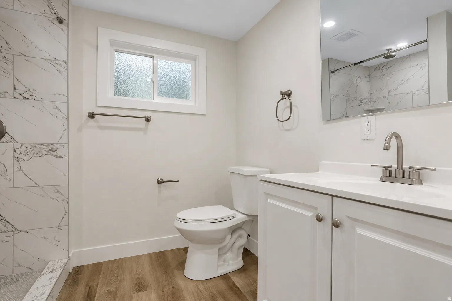 3/4 bathroom - Walk-in shower with marble-look tile, white vanity with integrated sink, wood-finish flooring, and a frosted window
