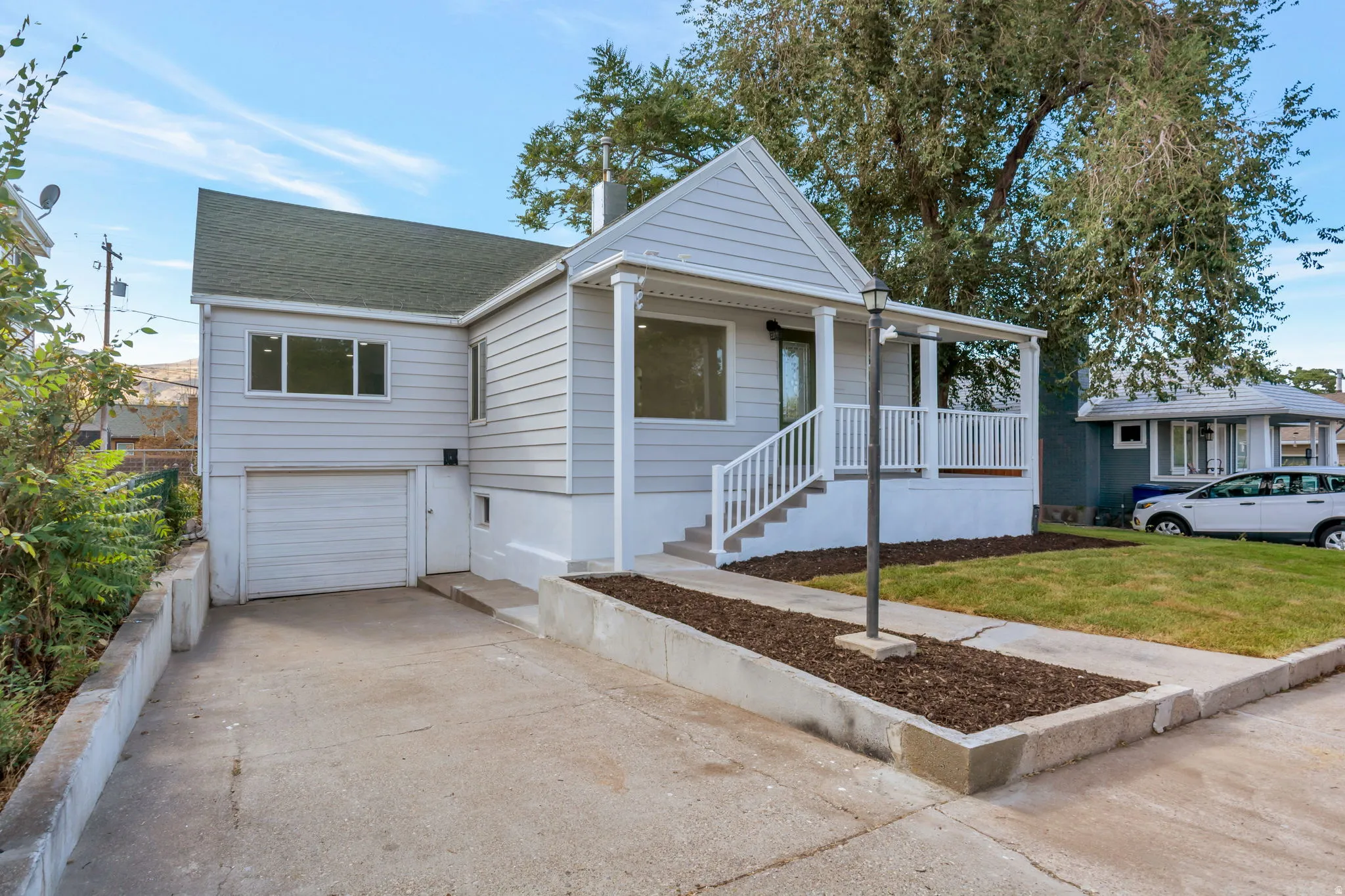 Light-toned siding exterior with a dark green roof, featuring a covered front porch with white railing and steps, an integrated garage, and a concrete driveway