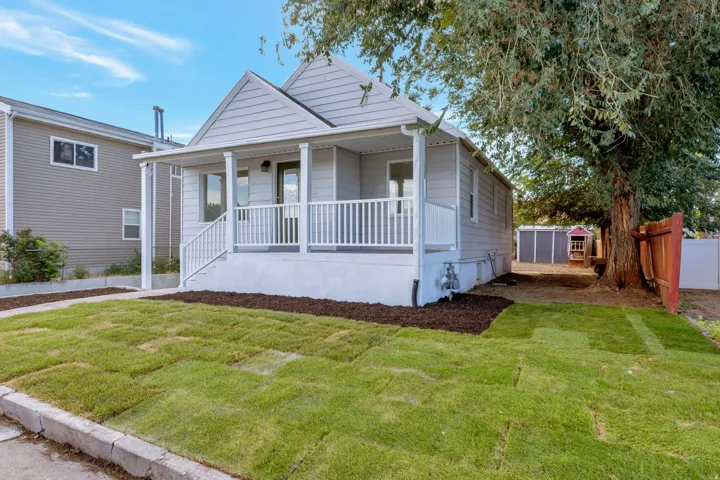Light gray siding exterior with a covered front porch featuring white railings, a front door, a mature tree, and a fenced yard