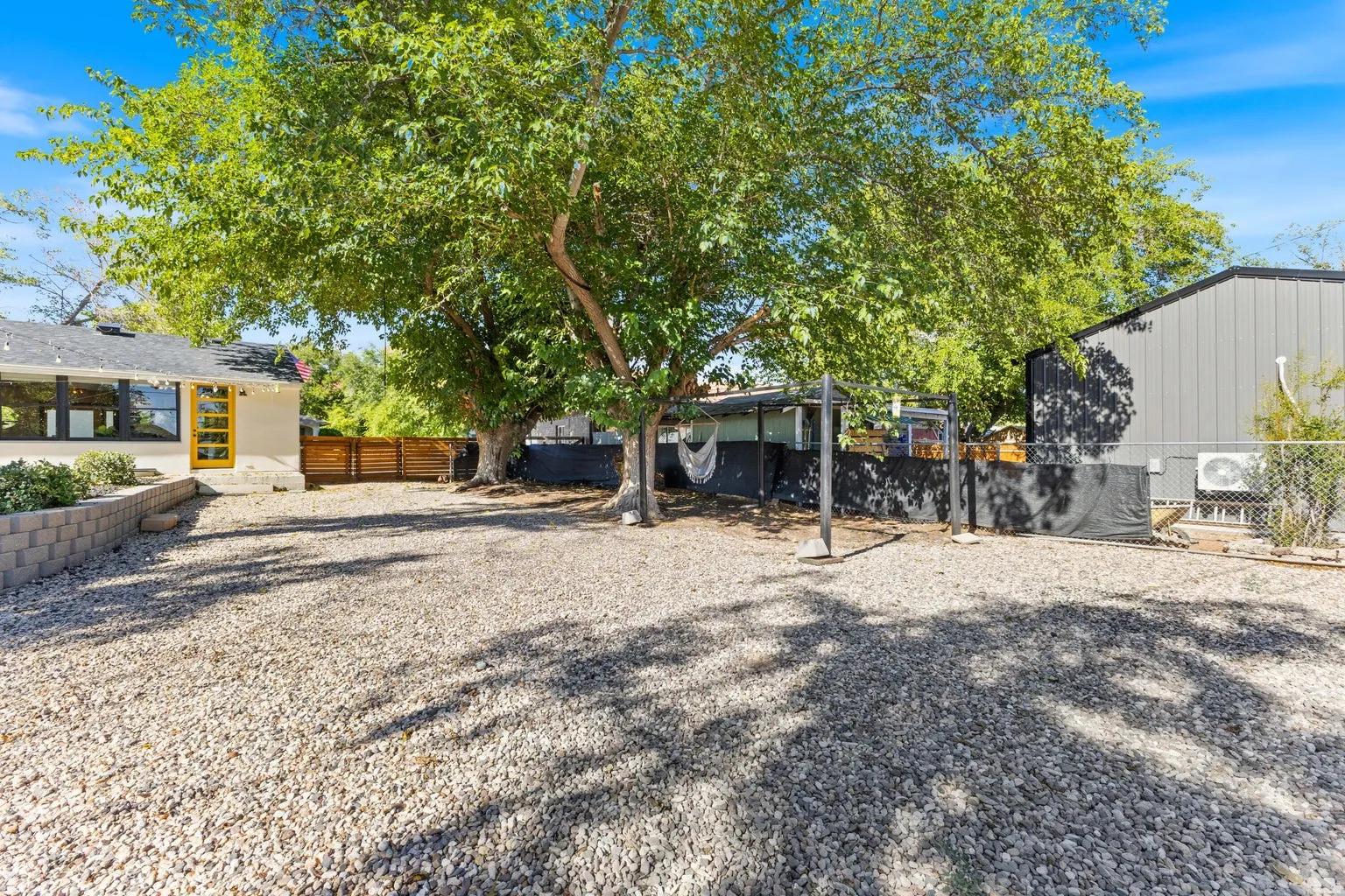 Expansive gravel yard featuring mature trees, a retaining wall, and a wood privacy fence