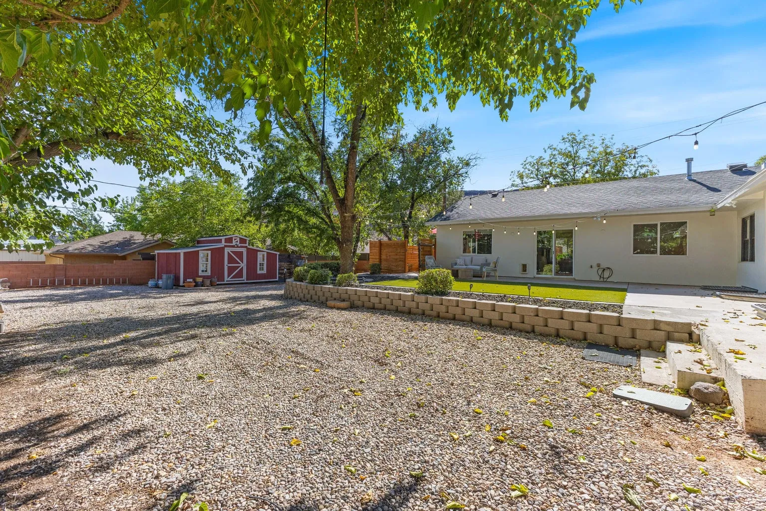 Expansive backyard featuring a gravel area, a red storage shed, a raised planter bed with block retaining wall, a concrete patio, and a section of artificial turf