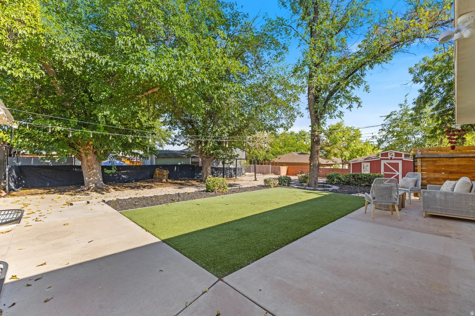 Spacious concrete patio with a detached shed, mature trees, and an artificial turf lawn