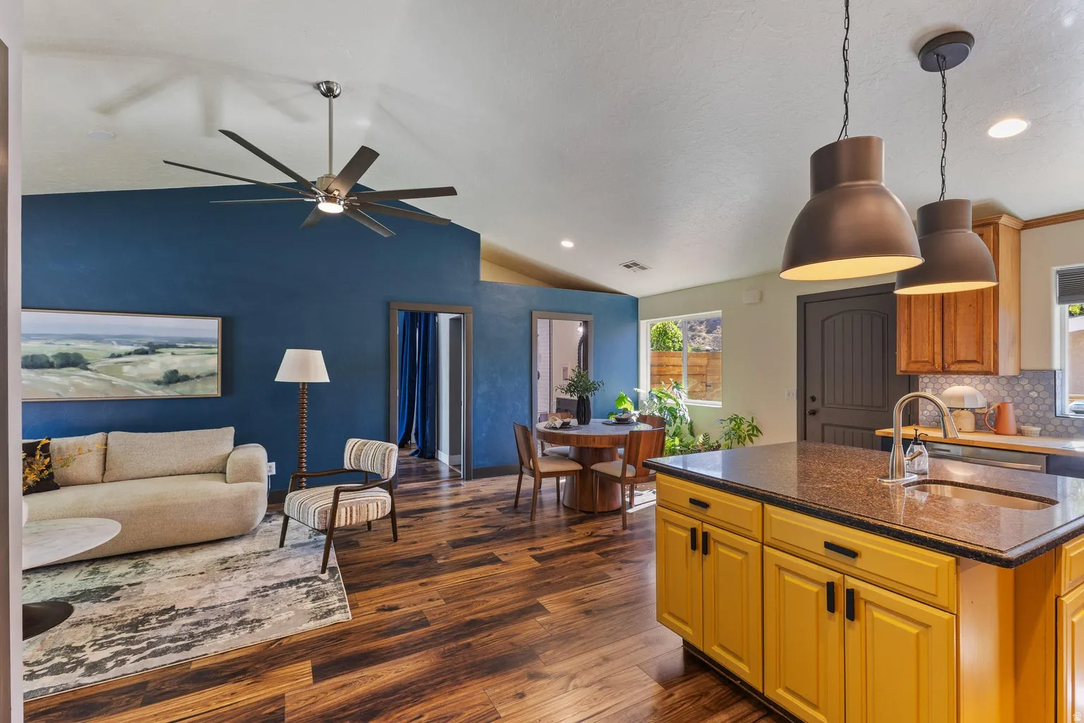 Vaulted ceiling with a large ceiling fan, wood-finish flooring, a kitchen island with a built-in sink, pendant lighting, and a backsplash