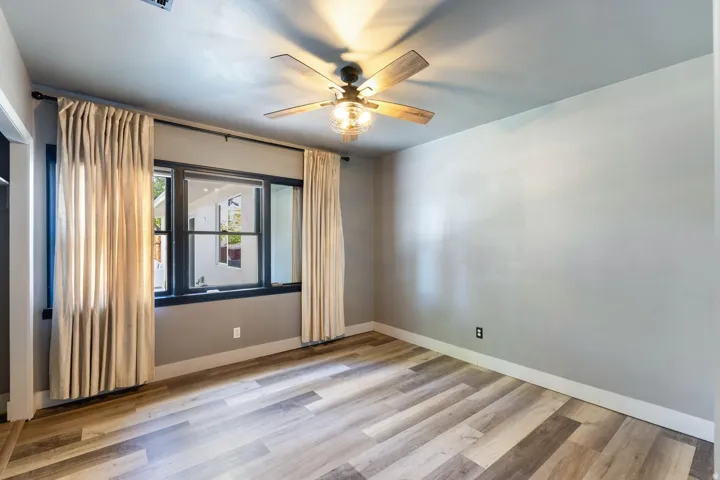 Spacious bedroom featuring hardwood flooring in warm, mixed tones, ceiling fan with integrated lighting, and large window with natural light and custom drapery