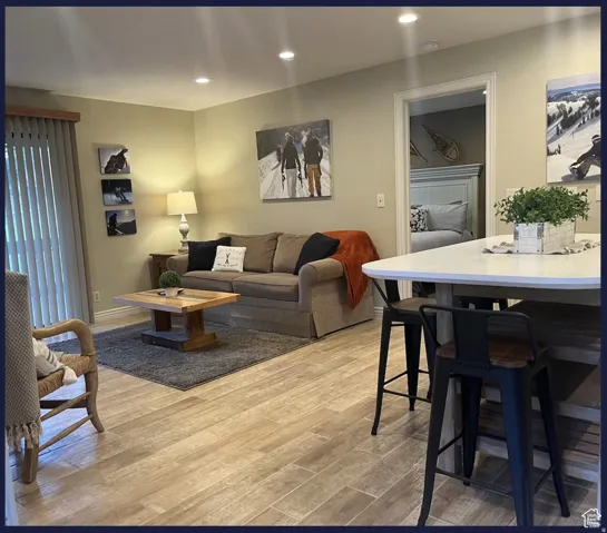 Recessed lighting throughout, wood-finish flooring, built-in breakfast bar with white countertop, and a sliding glass door with vertical blinds