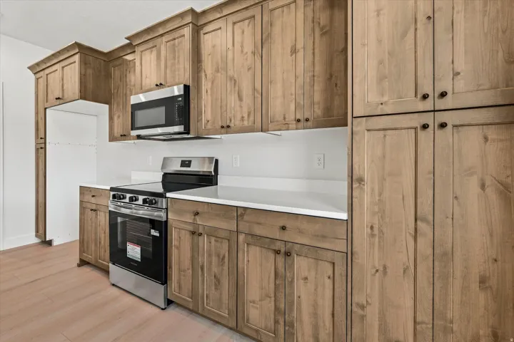 Wood-finish cabinetry, stainless steel microwave, electric range, white countertops, and light-toned wood-finish flooring