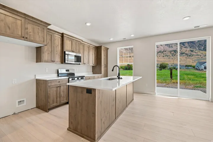 Open-concept kitchen with wood-finish cabinetry, white countertops, stainless steel appliances, and wood-finish flooring, featuring a large kitchen island with an integrated sink and a sliding glass door to the exterior