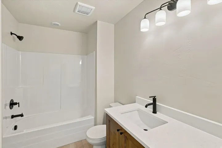 Bathroom featuring a white tub/shower combination with matte black fixtures, a single vanity with a white countertop and integrated sink, a wood-finish cabinet, and a four-light vanity fixture