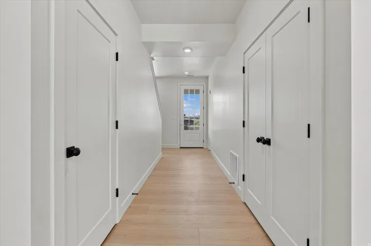 Hallway featuring light wood-finish flooring, white walls, multiple white doors with black hardware, and recessed lighting