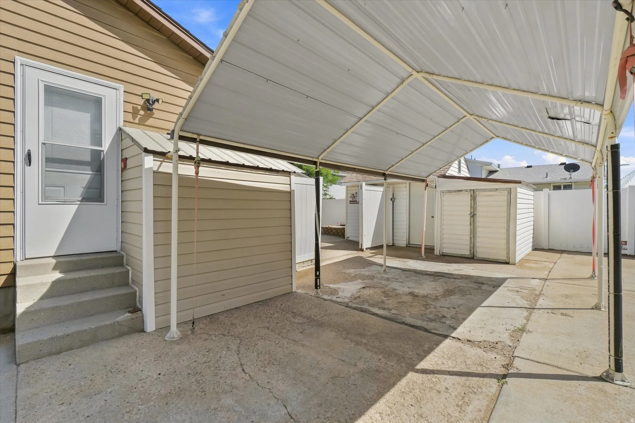 Covered patio with concrete flooring, multiple detached storage sheds, and a side entry door