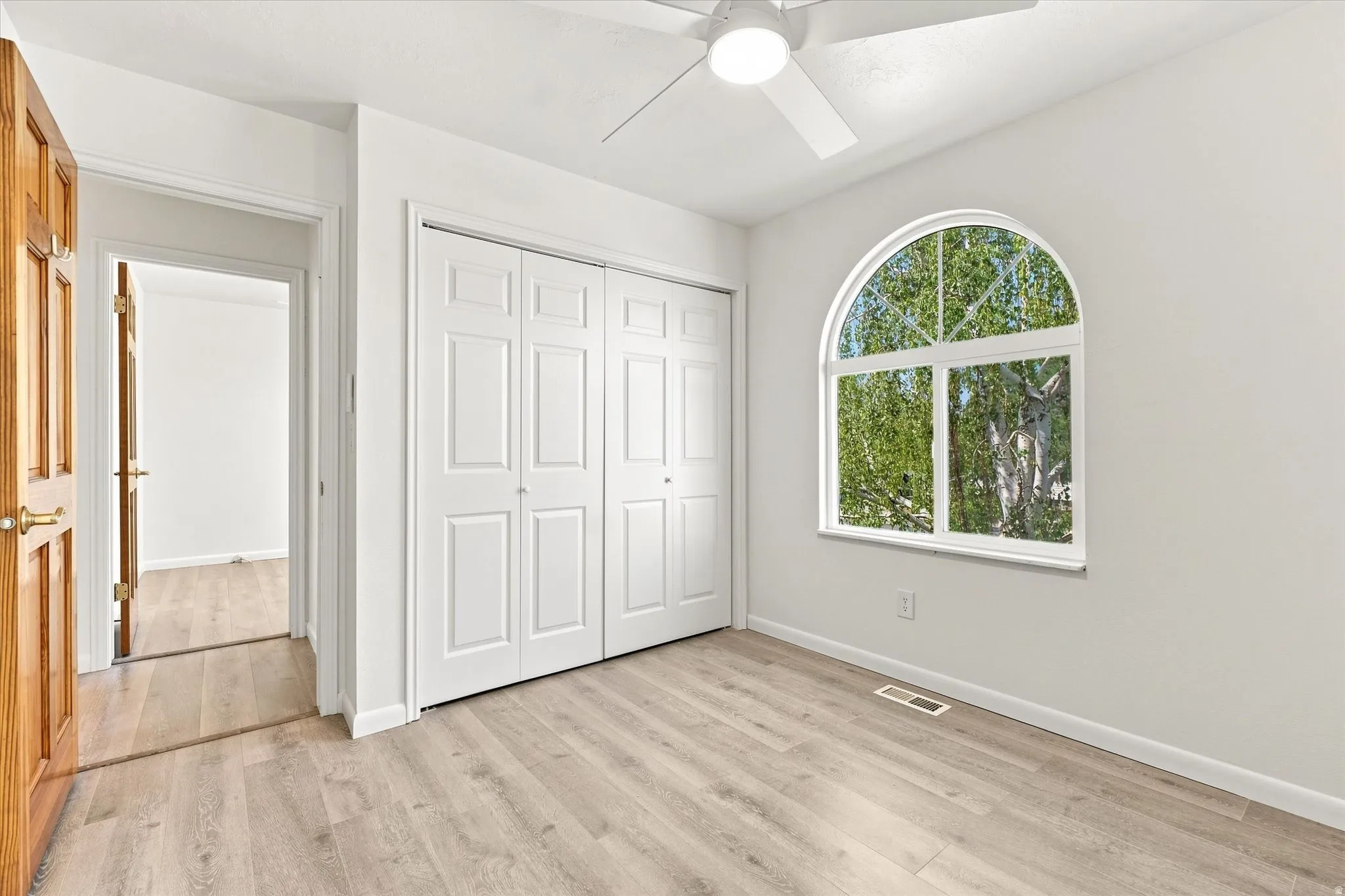 Arched window with exterior tree scenery, wood-finish flooring, white bi-fold closet doors, and a ceiling fan