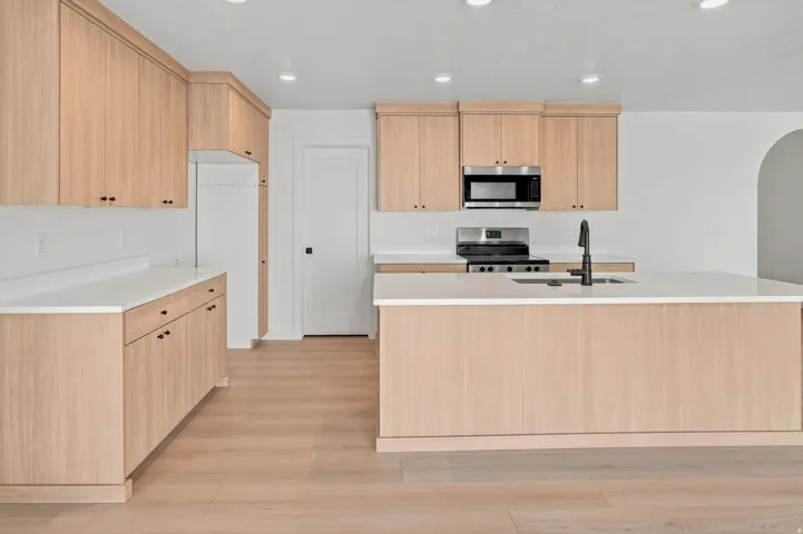 Light wood-finish cabinetry with dark hardware, white countertops, stainless steel appliances, and a central island with an integrated sink and matte black faucet