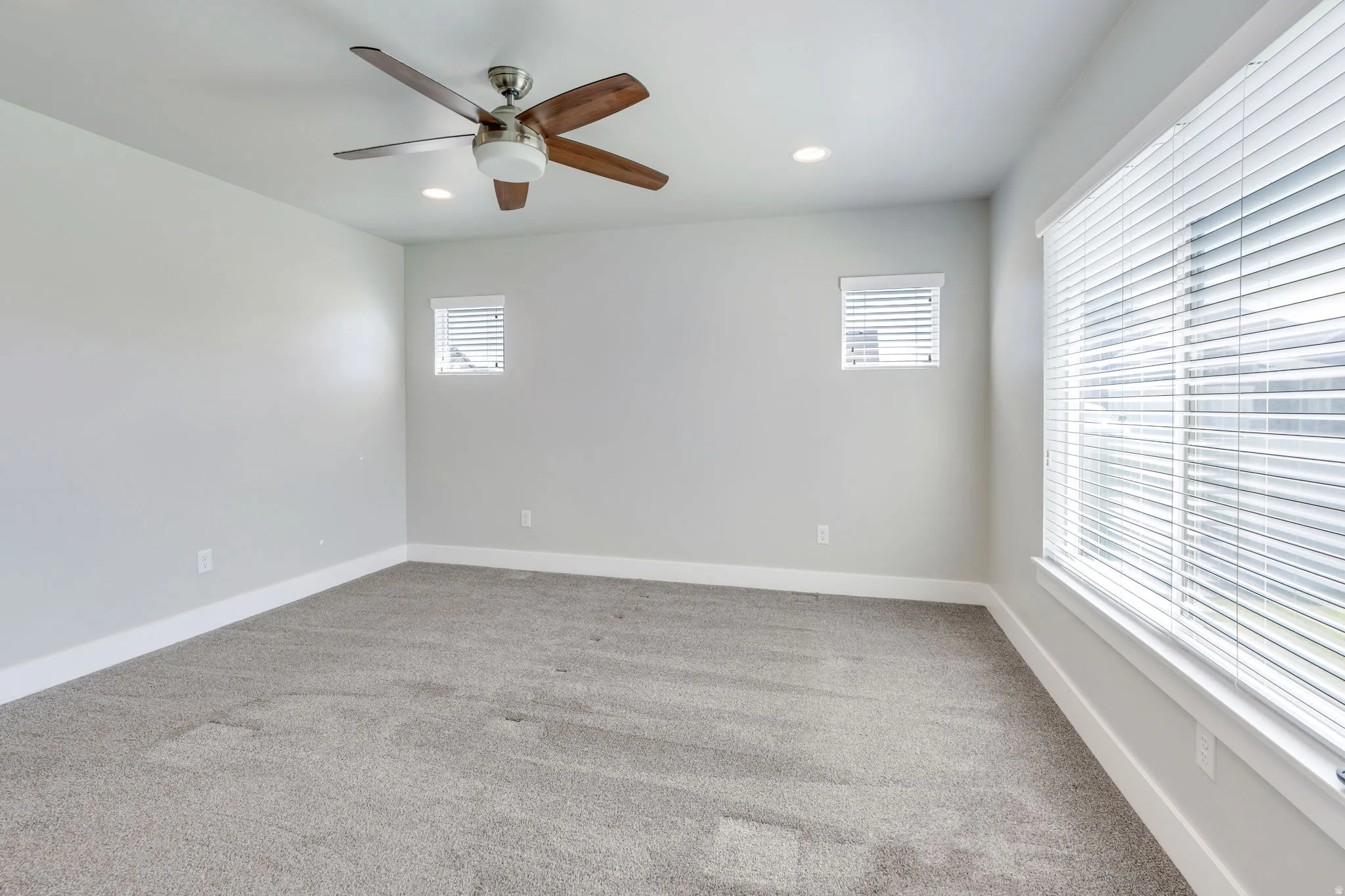 Recessed ceiling lighting, ceiling fan with wood-finish blades, multiple windows with blinds, and light gray wall paint