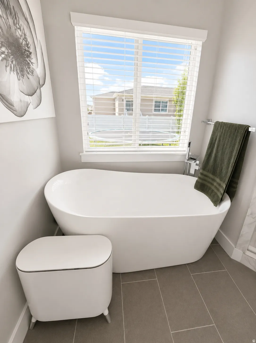 Freestanding soaking tub with a wall-mounted faucet, tile flooring, and a large window with blinds