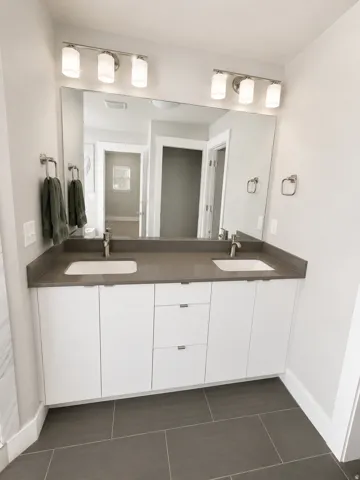 Dual vanity with integrated sinks and a dark-toned countertop, white cabinetry with silver-toned hardware, large wall-mounted mirror, and overhead lighting fixtures