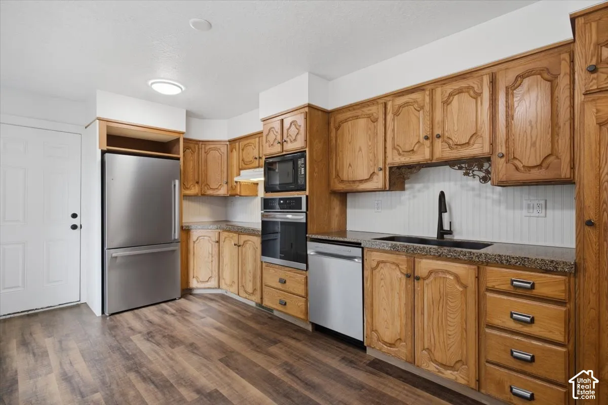 Kitchen featuring wood-finish cabinetry, stainless steel appliances, speckled countertops, a dark sink with a matte black faucet, and wood-finish flooring