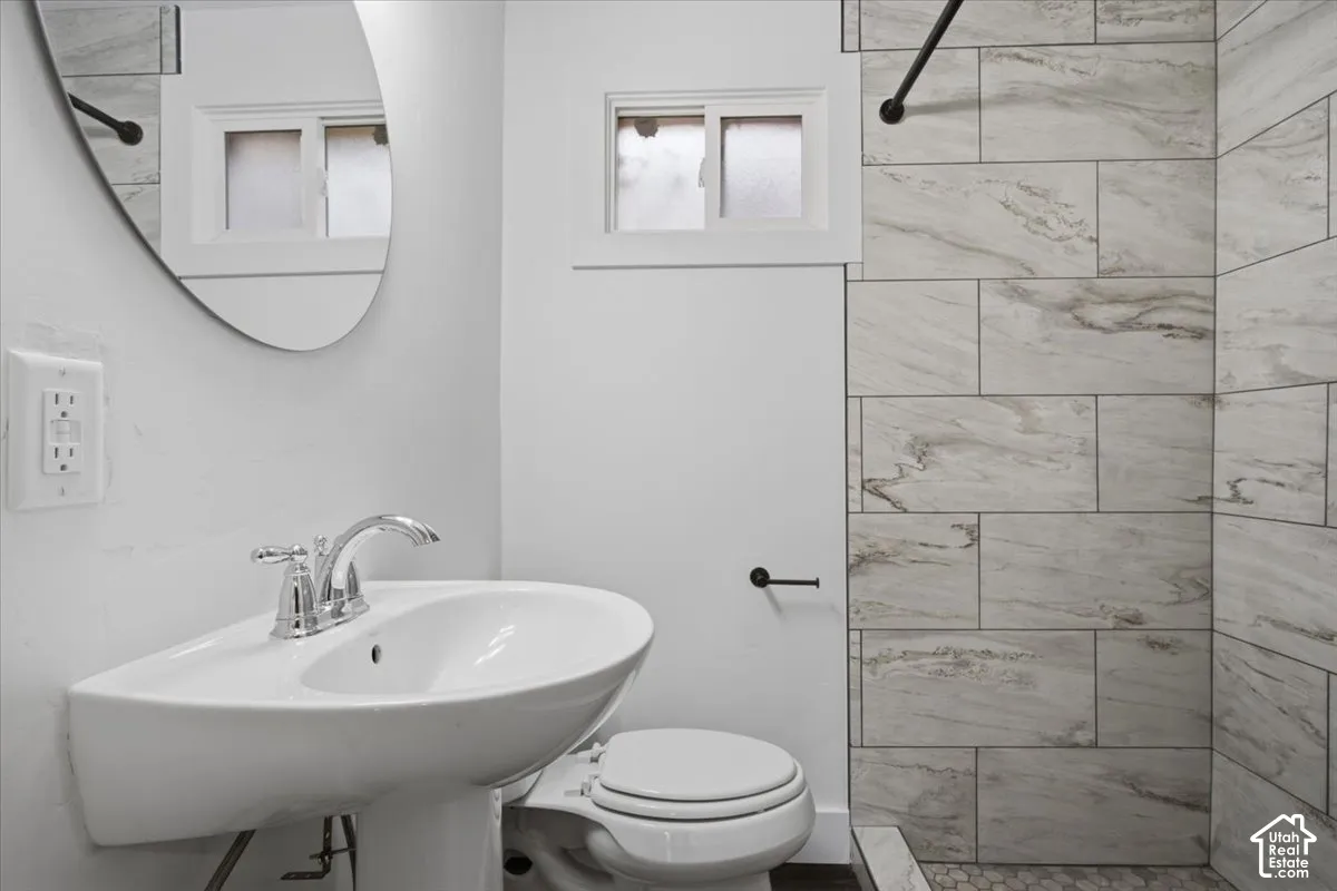 Pedestal sink with chrome fixtures, oval mirror, window with frosted glass, and shower with large format tile surround