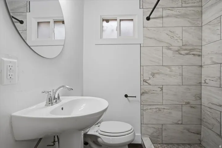 Pedestal sink with chrome fixtures, oval mirror, window with frosted glass, and shower with large format tile surround