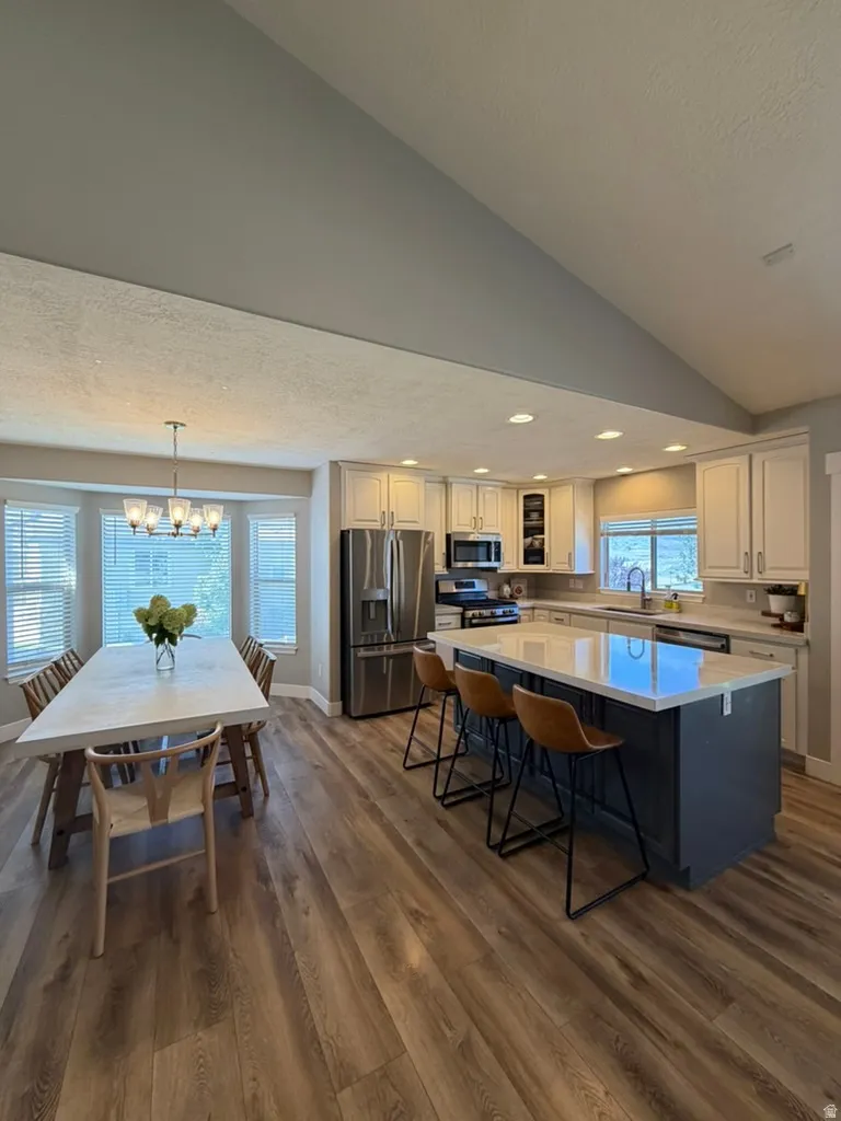 Open-concept kitchen and dining area featuring vaulted ceilings with recessed lighting, wood-finish flooring throughout, stainless steel appliances, blue-toned island with seating for multiple, cream cabinetry with dark countertops, and abundant natural l