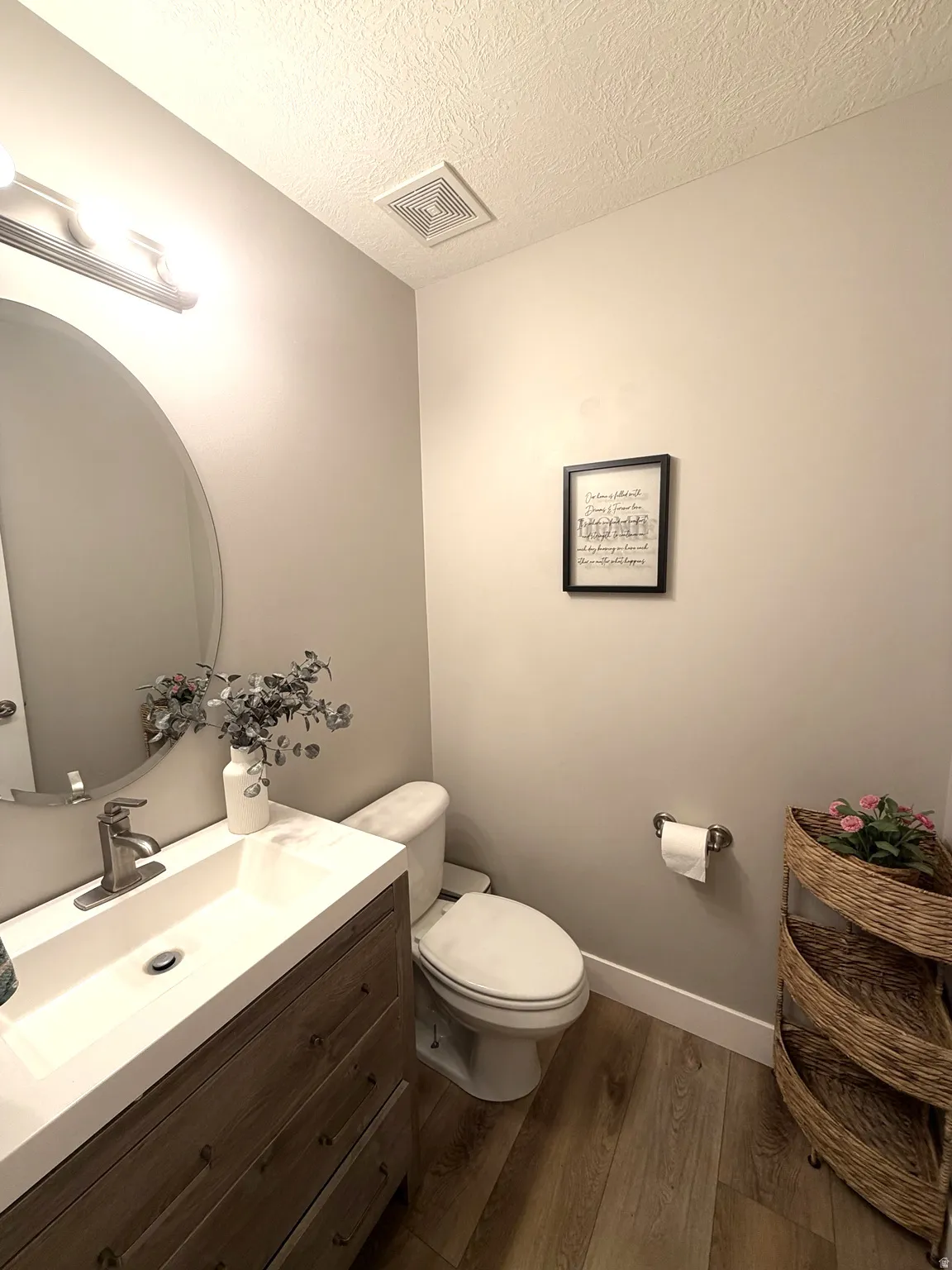 Wall-mounted vanity light fixture, oval mirror, wood-finish flooring, white toilet, and a vanity with an integrated sink
