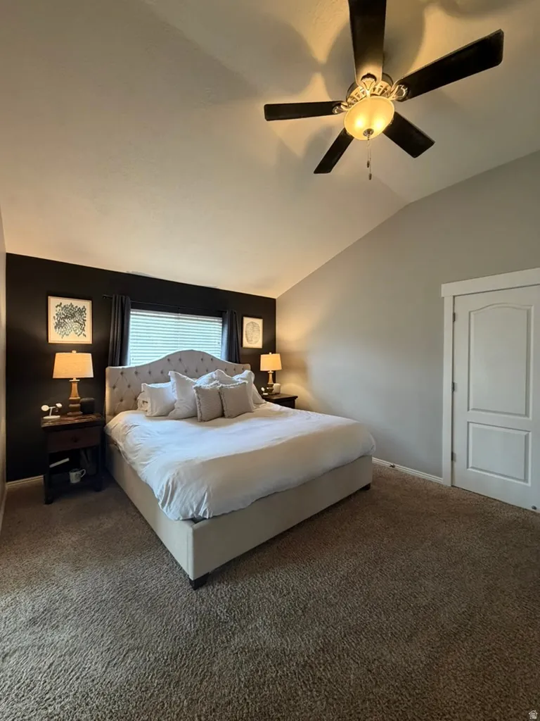 Master bedroom featuring a vaulted ceiling with ceiling fan and integrated lighting, neutral carpet flooring, dark wood-finish accent wall with built-in nightstands and reading lamps, upholstered bed frame, and an ensuite door providing private access to