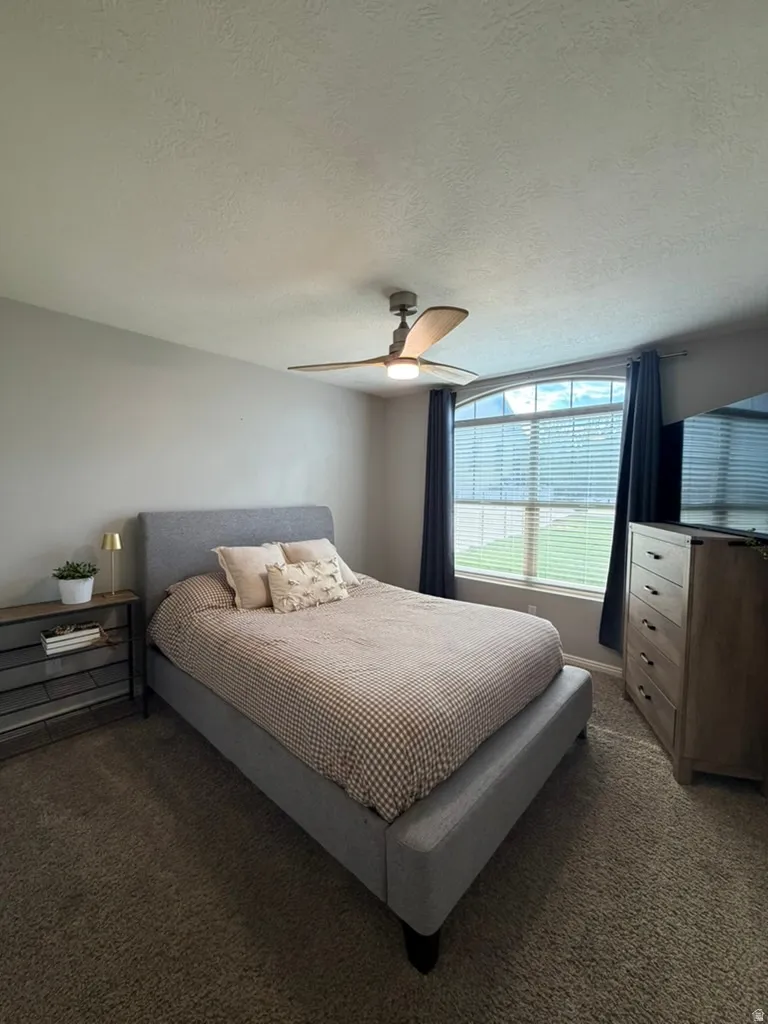 Bright secondary bedroom featuring a gray upholstered bed frame, dual nightstands with integrated lighting, wood-finish dresser, ceiling fan with integrated light fixture, and large multi-pane windows with blackout window treatments