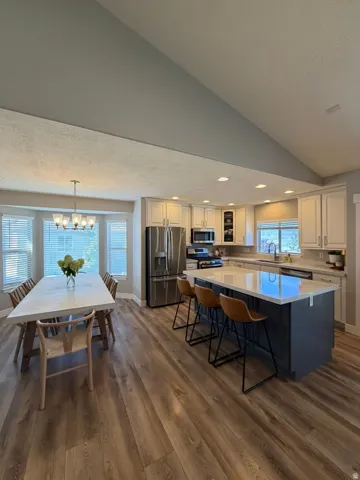 Open-concept kitchen and dining area featuring vaulted ceilings with recessed lighting, wood-finish flooring throughout, stainless steel appliances, blue-toned island with seating for multiple, cream cabinetry with dark countertops, and abundant natural l