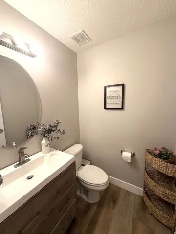 Wall-mounted vanity light fixture, oval mirror, wood-finish flooring, white toilet, and a vanity with an integrated sink