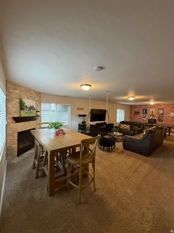 Open-concept living area featuring a wood-finish dining table with seating for six, exposed brick accent walls, a gas fireplace with brick surround, recessed ceiling lighting, and carpeted flooring throughout