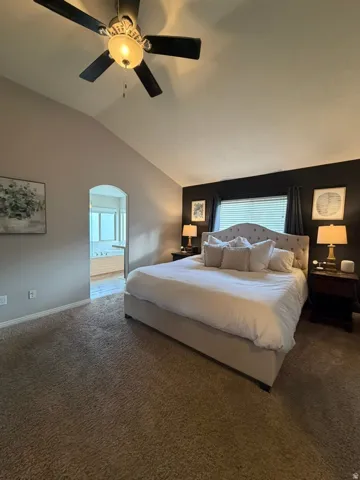 Master bedroom suite with vaulted ceiling, ceiling fan with integrated lighting, carpeted flooring throughout, and an ensuite bathroom visible through the arched doorway