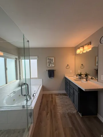 Spa-inspired primary bathroom featuring a freestanding soaking tub with glass enclosure, dual vanity with wood-finish cabinetry and quartz countertops, wood-finish flooring, and contemporary lighting fixtures
