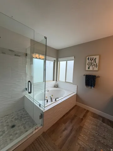Spa-inspired primary bathroom featuring a frameless glass-enclosed walk-in shower with subway tile surround and mosaic floor, freestanding soaking tub with deck-mounted fixtures and windows overlooking the landscape, wood-finish flooring, and warm neutral