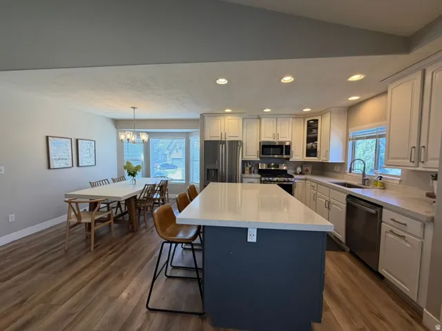 Open-concept kitchen and dining area featuring a blue-painted island with waterfall-edge countertop, stainless steel appliances, cream cabinetry with brushed nickel hardware, recessed lighting, wood-finish flooring throughout, and multiple windows providi