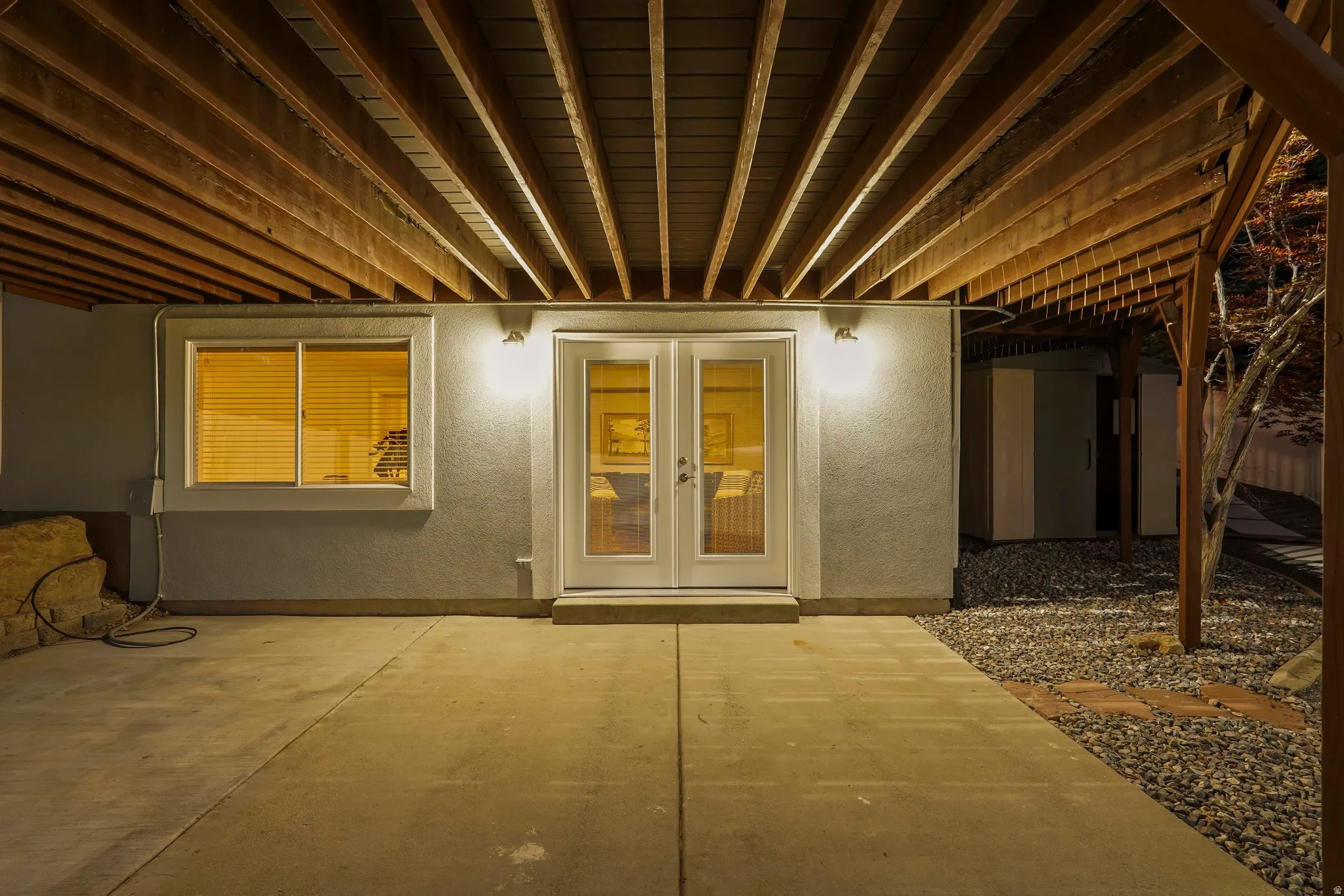 Covered patio with concrete flooring, exposed wood beam ceiling, white French doors, and exterior lighting