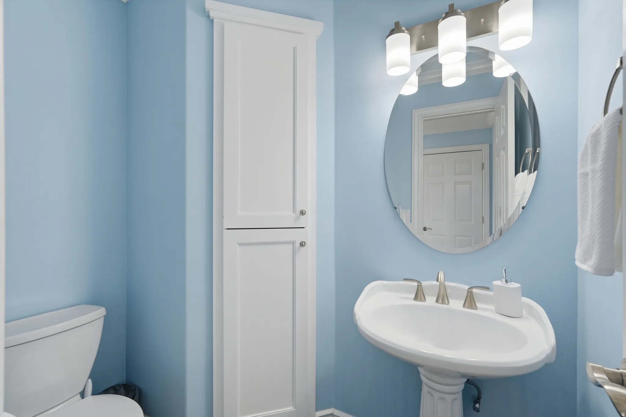 Pedestal sink, wall-mounted oval mirror, three-light vanity fixture, floor-to-ceiling storage cabinetry, and commode